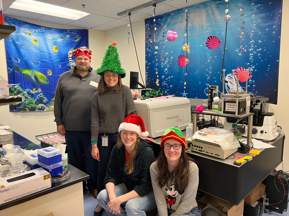 From left to right, Vizgen engineer Nick wearing and elf hat, Dr. Christina Baer in a christmas tree hat, Jill McConnell program manager in a santa hat, Molly Martin post doc in an elf hat all in front of the new Merscope Ultra. 