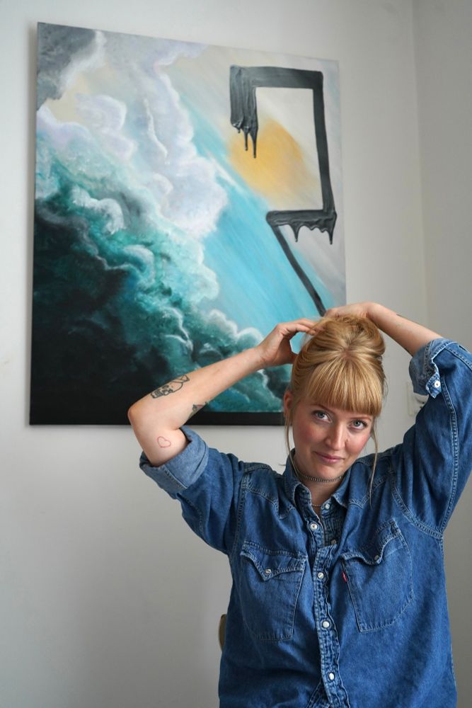 Blonde woman sitting in front of her painting. Putting her hair up - she’s ready to work on some new art pieces.