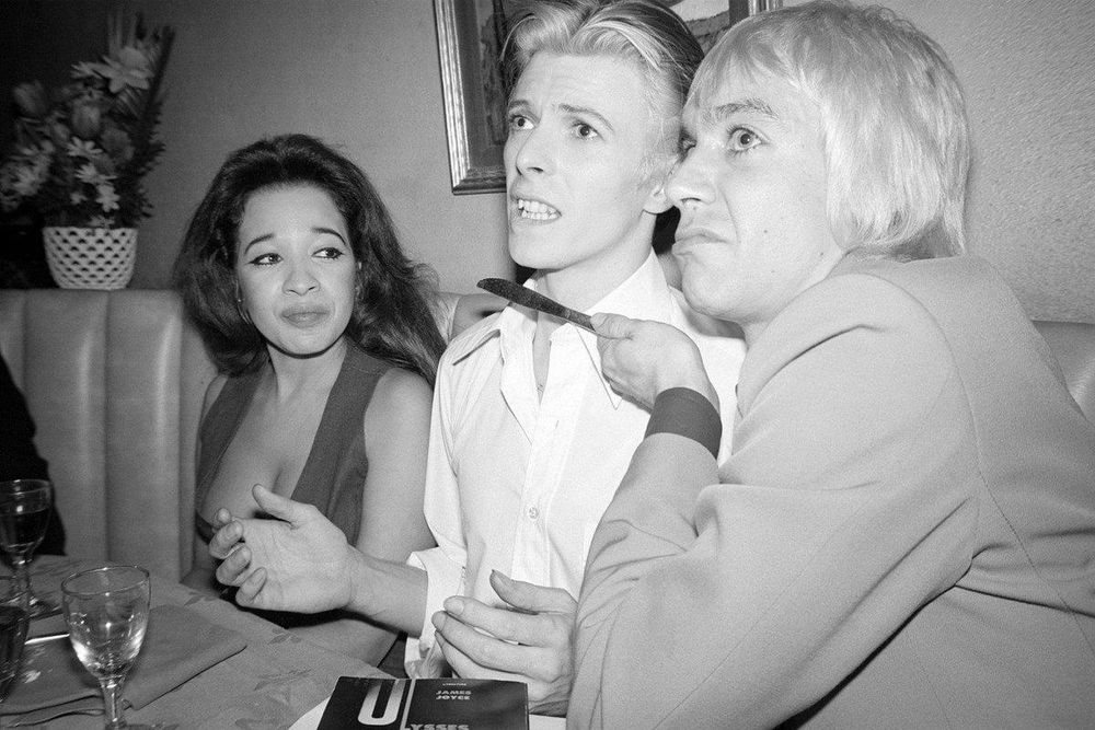 Iggy Pop holds a butter knife (meaning not sharp at all) to David Bowie’s neck and makes a nasty face at someone off camera threateningly. David Bowie throws his arms up with an expression that says “Oh darn, why is it always me?” Meanwhile Ronnie Spector is watching & visibly straining trying not to laugh at the whole thing. I do not have context, I think the dudes were just being silly.