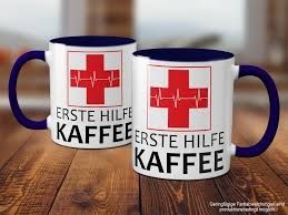 Zwei weiße Kaffeetassen mit dunkelblauem Henkel und Innenfarbe stehen auf einer Holzoberfläche. Auf jeder Tasse ist ein rotes Kreuz mit einem EKG-Linienmuster in der Mitte abgebildet. Darunter steht in Großbuchstaben der Text 'ERSTE HILFE KAFFEE'.
