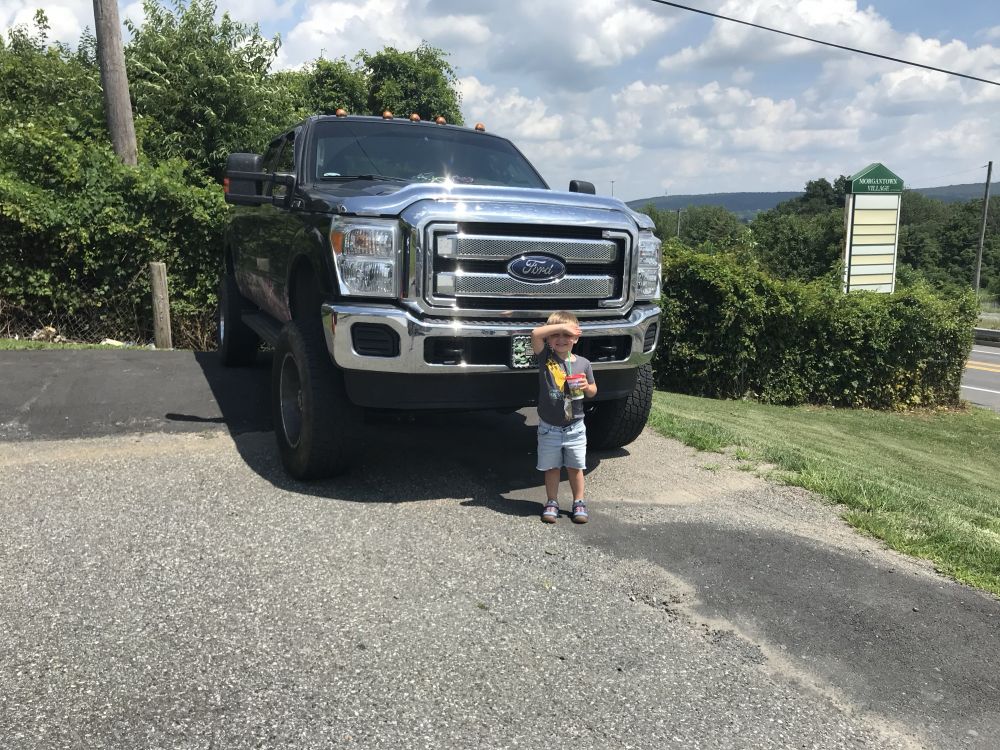 A small child stands in front of and is dwarfed by the grill of an oversized Ford pickup truck.