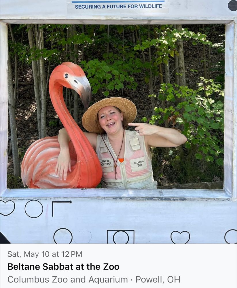 A person in a zookeeper vest and a sun hat hugs a flamingo statue and laughs