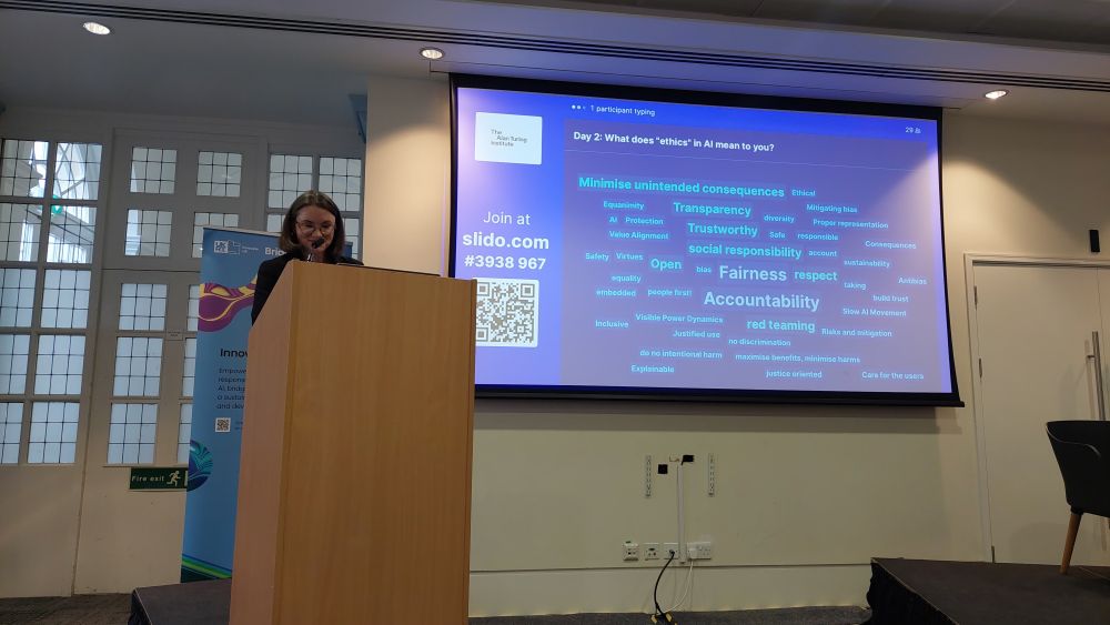 Arielle, a white British Woman, with short black hair, rimmed spectacles and black dress, is standing on the podium and looking at her laptop which is projected behind her, showing many terms that participants have shared about what they mean by ethics. Top 5 terms are accountability, fairness, trustworthy, transparency and social responsibility.