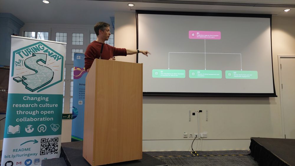 Chris, a short haired white British man, wearing a maroon sweater and is standing on the podium, next to The Turing Way banner. He is pointing on the screen that shows a workflow example of what kinds of question can be asked in ensuring assurance of AI tool/product.