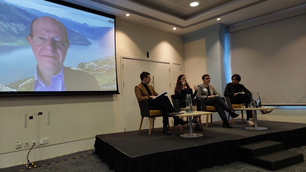 Four people sitting on the stage and another joining virtually.