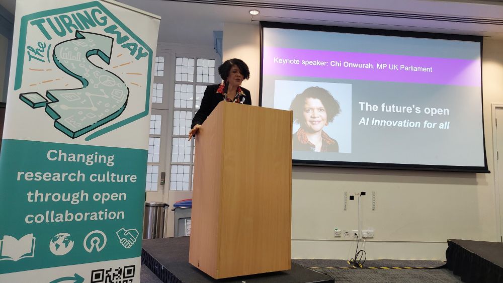 Chi Onuwara, a black woman, is standing on the podium and giving a keynote. She is a tall person, with short curly hair. On her right is a big Turing Way banner with logo on green saying "chaing research culture through open collaboration". On her left is a screen projecting a slide with her name and talks title "the Future’s Open- AI innovation for all".