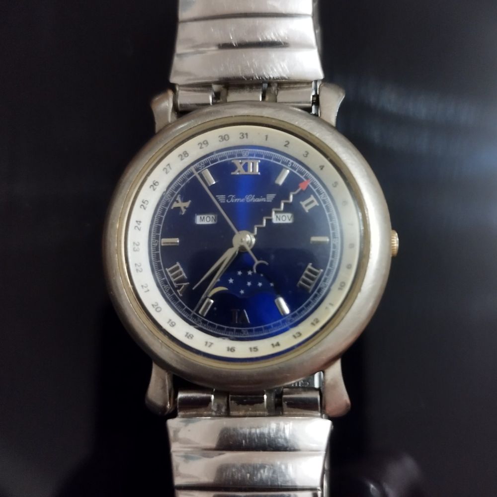 close up photo of a silver, round "Time Chain" wrist watch with a metallic dark blue face and roman numbers.
it has 4 hands, one of which is a zig zag with a red arrow tip pointing to the day of the month on the surrounding ring numbered 1 through 31.
the weekday and month are displayed in abbreviated form (MON and NOV).
a small rotating dial decorated with tiny five-pointed stars indicates the crescent stage of the moon.