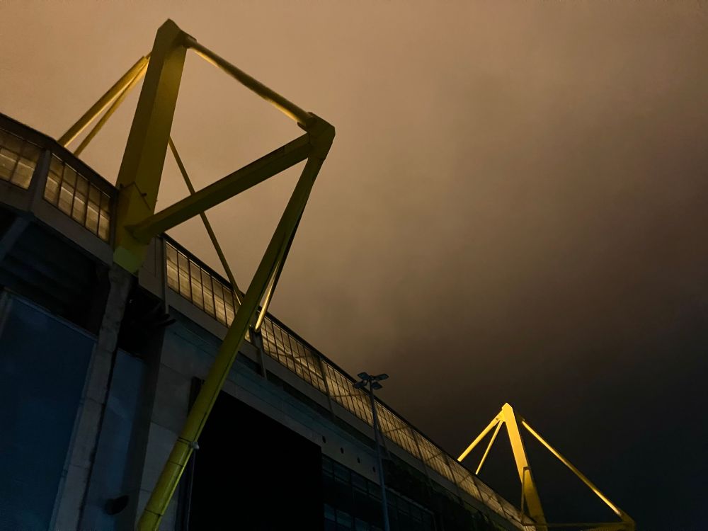 Zwei Pylonen des Westfalenstadions im dunstigen Nachthimmel. Das Licht, welches sie anstrahlt und die Nacht zeichnen sich als Schattenspiel auf ihrer Oberfläche ab. Während der vordere sich dunkel vor dem beleuchteten Hintergrund abhebt, wird der hintere von Scheinwerfern angestrahlt und hebt sich hell vor der dunklen Nacht ab. 