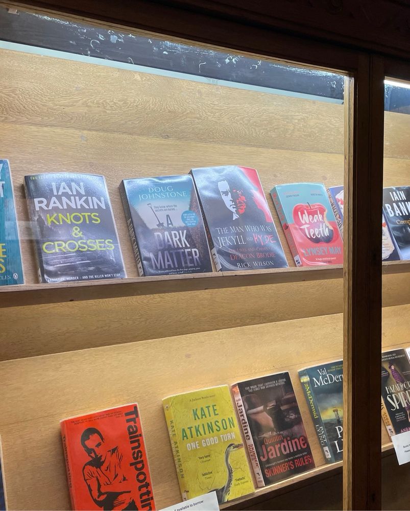 Gently lit bookshelves in a wood and glass cabinet, with books facing outwards. Including weak teeth, train spotting and gear no evil