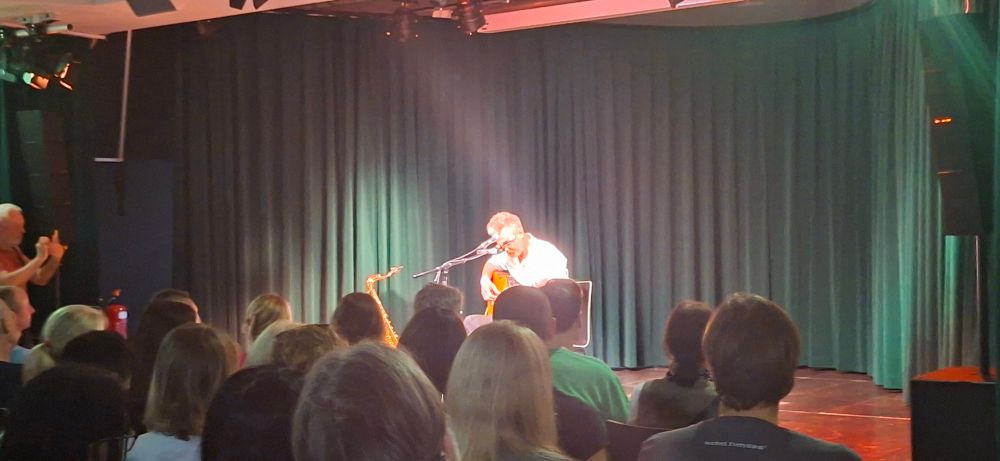 Matt Elliott auf einer kleinen Bühne mit Gitarre am Schoß, vor ihm Mikrofonständer. Publikum ist im Vordergrund sichtbar.