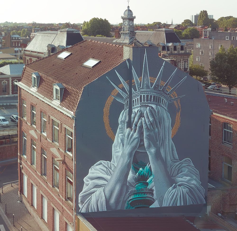 Mural by JDL Street Art in Roubaix, Lille, France, depicting the Statue of Liberty covering her face with both hands in a gesture of shame. The figure is painted in shades of grey with teal accents on the torch and crown, set against a muted background with a golden halo-like circle. The artwork spans the side of a tall brick building, contrasting modern street art with historic European architecture in the surrounding cityscape.