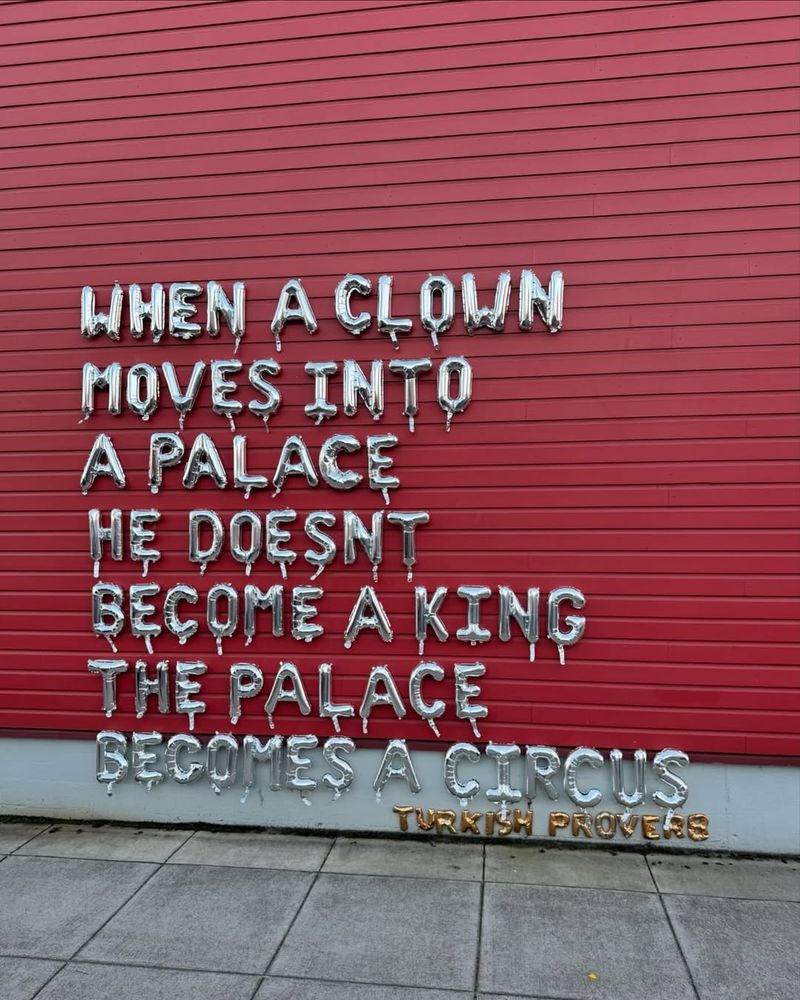 A striking street art installation by BLCKSMTH, created using metallic silver balloons spelling out a well-known Turkish proverb: "WHEN A CLOWN MOVES INTO A PALACE, HE DOESN'T BECOME A KING. THE PALACE BECOMES A CIRCUS." The text is mounted on a red wall, with "TURKISH PROVERB" highlighted in gold balloons at the bottom. The slightly deflated balloons and dripping effect enhance the visual impact of the message. Created in response to Donald Trump’s return to office in 2025, the piece transforms public space into a striking political statement.