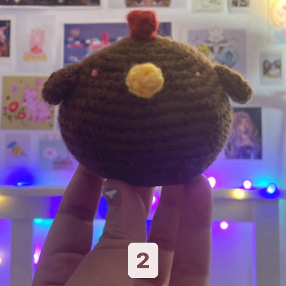 Crochet plushie of a brown fat chicken with pink cheeks and yellow beak being held by a pink hand.
In the background there are pink, yellow and blue lights and a lot of posters