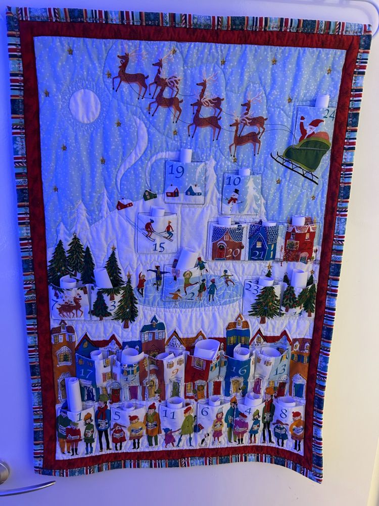 A handmade advent calendar approximately 20 inches by 24. The image is of Santa's sleigh flying over a snowy village. Small scrolls of paper can be seen in the pockets for the 1st to 24th December.