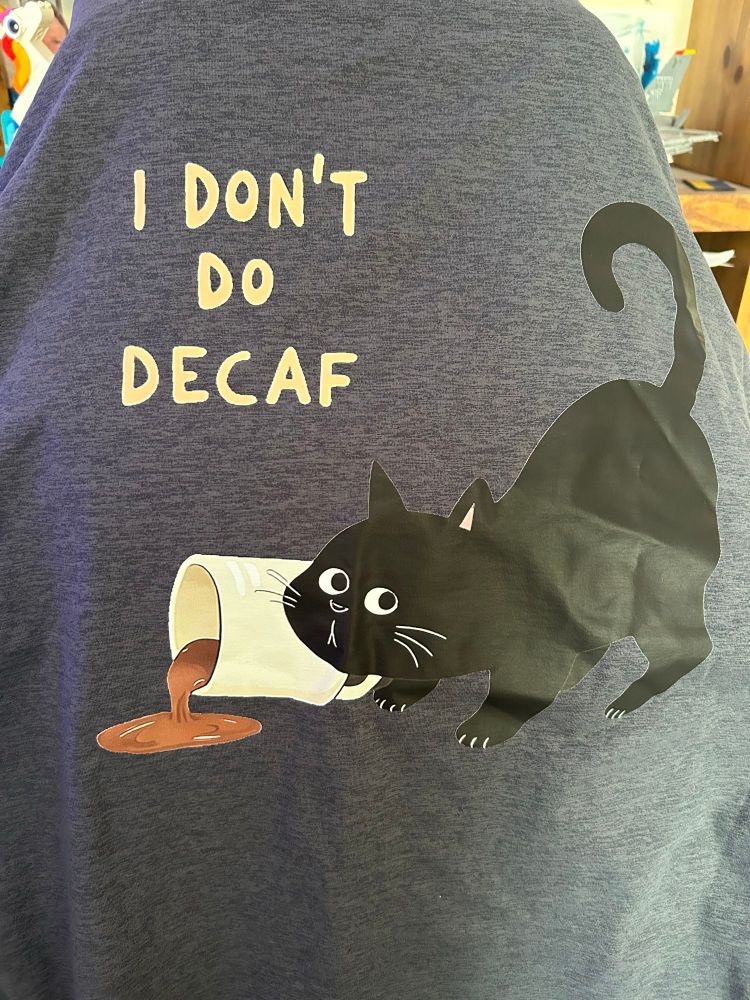 A dark grey tee shirt draped over an office chair. A cartoon black cat is crouched, tail high, by a pushed over white mug of coffee. Text reads I don’t do decaf. 