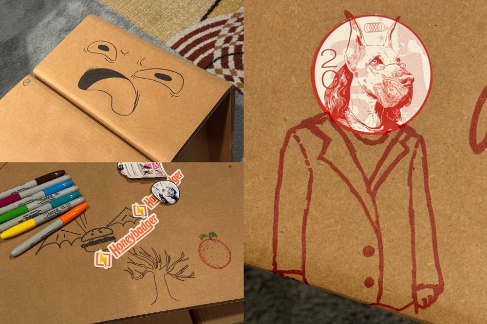 Detail shot of Honeybadger's interactive cardboard furniture showing attendee creativity with hand-drawn artwork including a smiling face, a tree, and a detailed figure drawing of a person with an animal head (a great dane). Sharpie markers and Honeybadger stickers are scattered around, encouraging conference attendees to personalize the space.