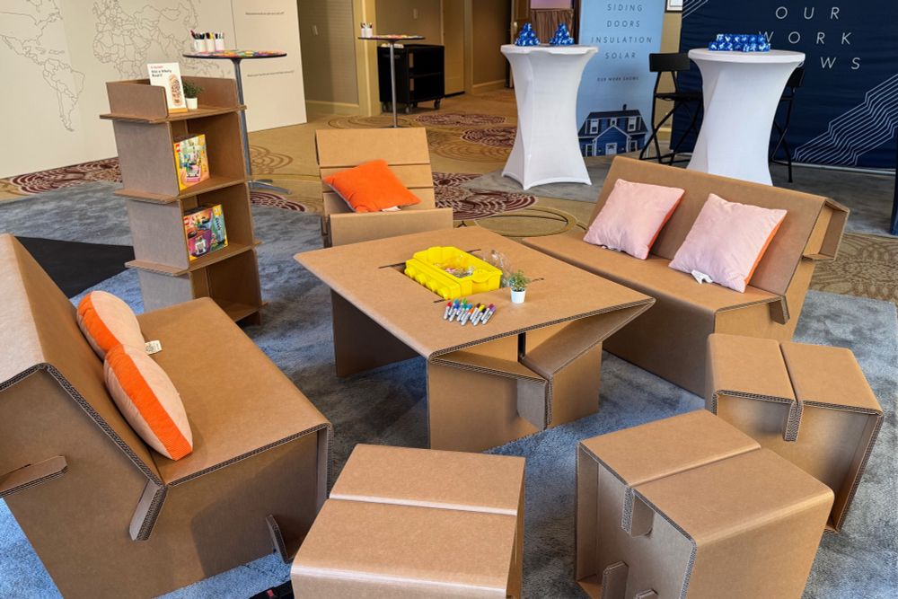 Honeybadger's cardboard furniture lounge setup at a conference, featuring fully functional cardboard chairs, sofas, coffee tables, and bookshelves with colorful pillows, markers for interactive drawing.