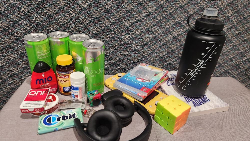 Photo shows 4 redbulls, mio drink enhancer, melatonin, ibuprofen, on! nicotine pouches, zyn nicotine pouches, headphones, earbuds, orbit gum, dramamine, two fidget toys, a rubik's cube, switch lite, 4 switch games, a book, and a large waterbottle