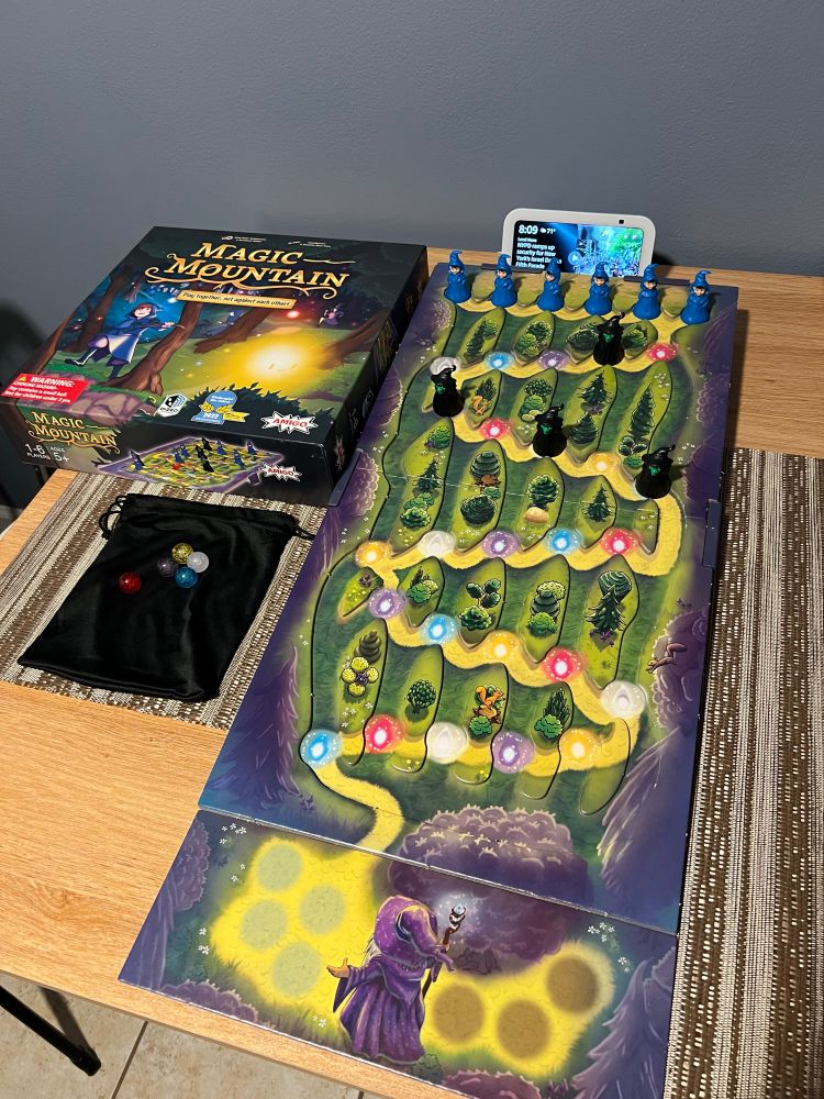 Photo if Magic Mountain boardgame box cover art, colored marbles over black bag, and play board with path.