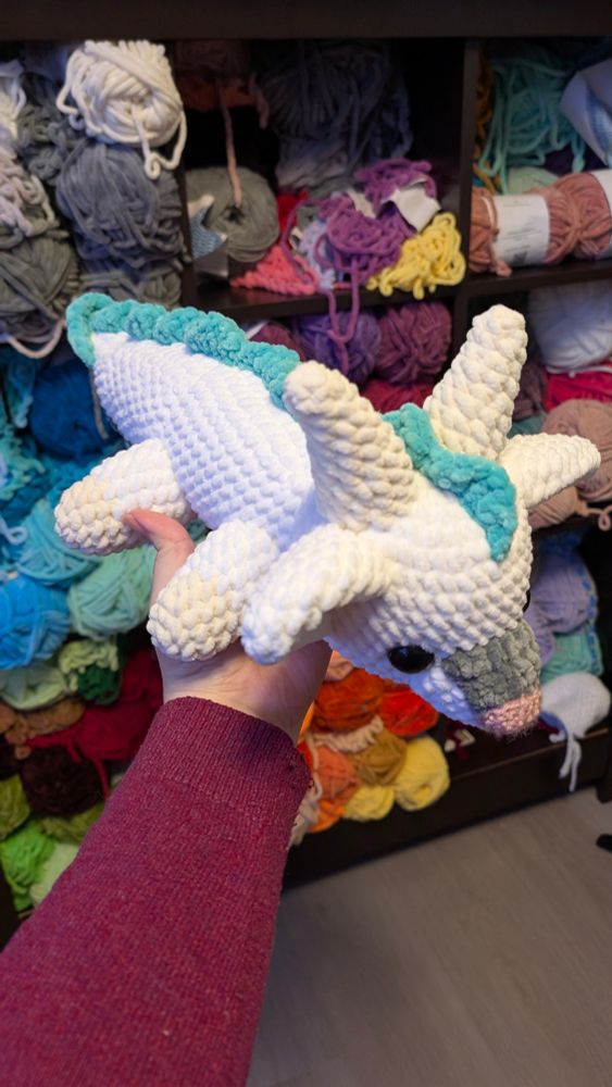 A crochet chibi version of the dragon form of Haku from the animated film Spirited Away. He is facing down and to the right slightly, allowing his body to be seen behind him. He has a white body, a grey patch on his snout, a pink nose, cream coloured horns, and an icy green coloured man going from head to tail. Behind him is a shelf filled with colourful yarn