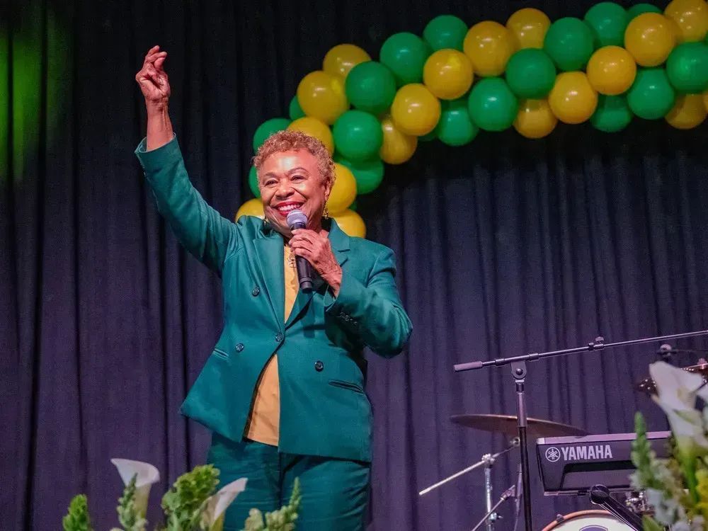 Barbara Lee, new mayor of Oakland, standing on a small stage in an green suit with yellow shirt, with green and yellow balloons behind her