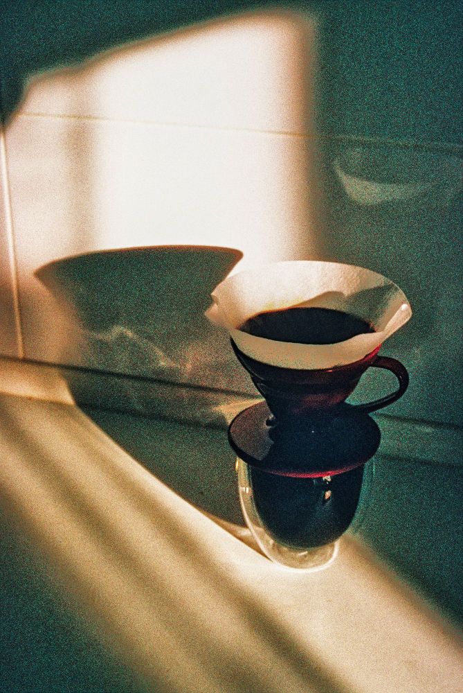 The image shows a coffee-making setup, specifically a pour-over coffee dripper placed on top of a clear glass. The dripper appears to be a dark color, possibly deep red, and is fitted with a paper filter containing coffee grounds. The scene is lit with warm, natural light that casts soft shadows and highlights on the wall behind the setup. The lighting creates a cozy, atmospheric feel, with light streaks and the shadow of the dripper contributing to the overall aesthetic. The image has a vintage or film-like quality, with some grain visible, adding to its nostalgic appeal.