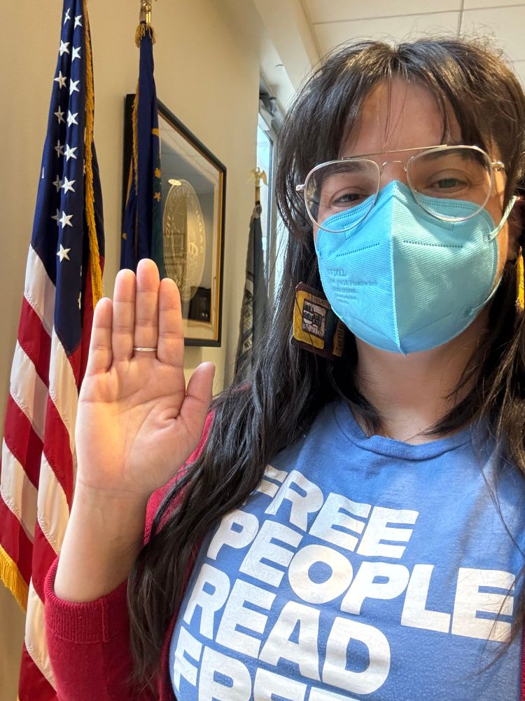Latina librarian wearing a face mask holding her right hand up in front of an American flag