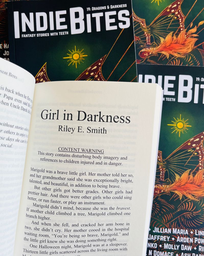 A stack of paperback copies of Indie Bites 19 with a cover illustrated by Arden Powell featuring a dragon. The top copy is open to the title page of Riley’s story.