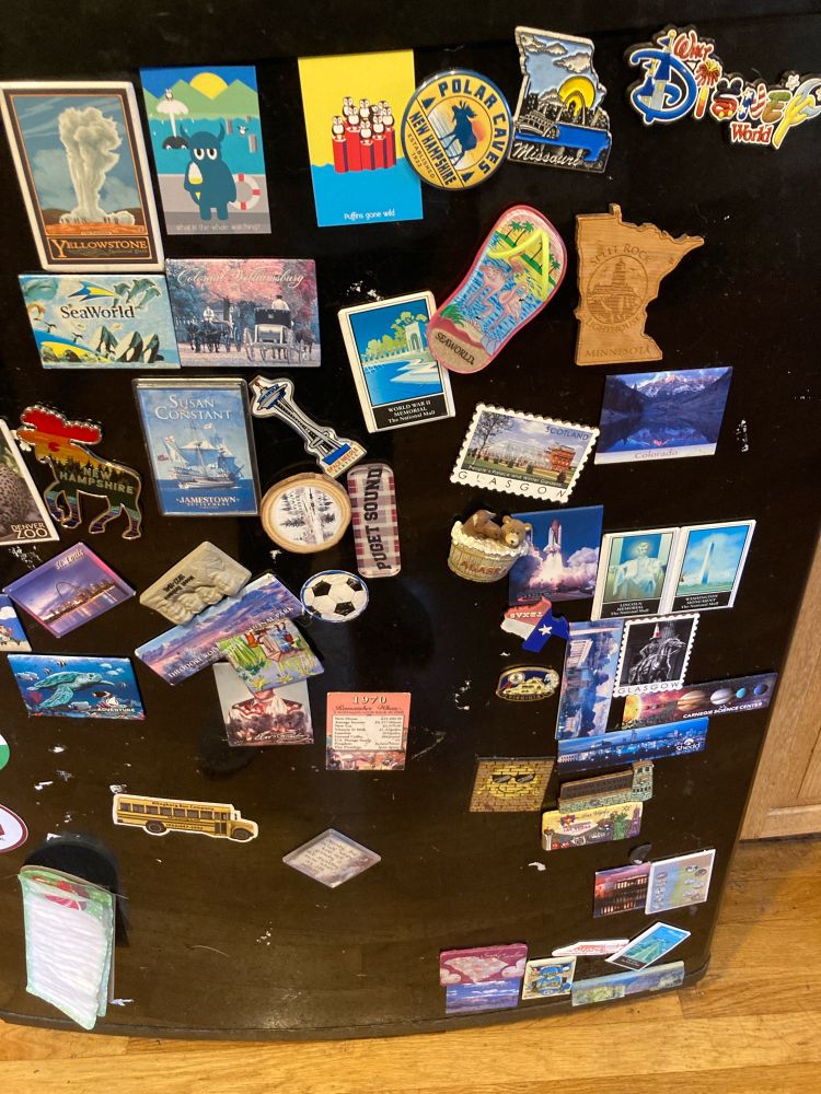 Chaotic picture of a black fridge with a lot of magnets haphazardly scattered. Magnets are from places around the US and world. 