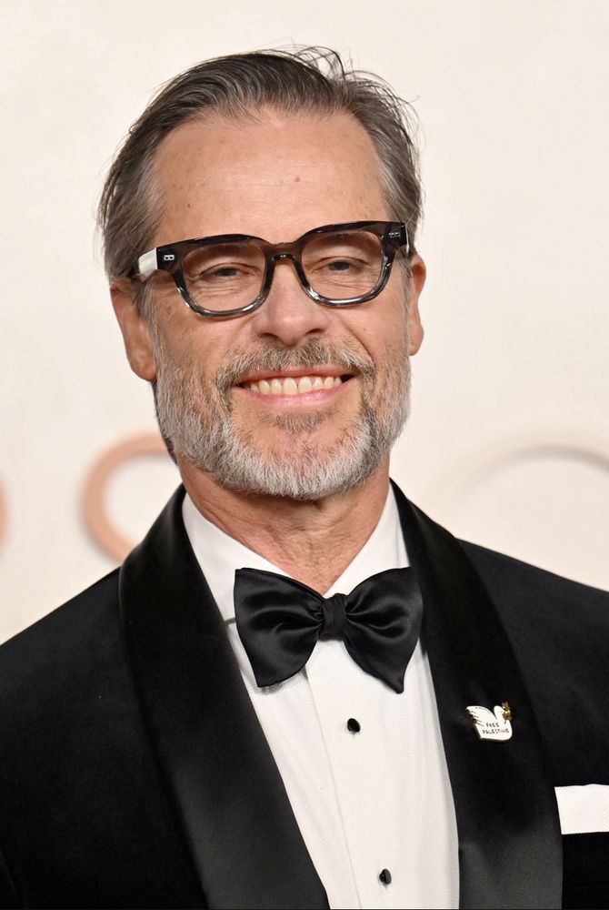 Guy Pearce wearing a Free Palestine pin on his jacket lapel. The pin is of a white dove with an olive branch in its beak, with the words 'Free Palestine' on its body. 