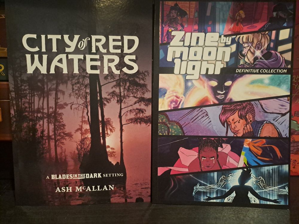 An image of physical copies of the TTRPG supplements 'City of Red Waters' for Blades In The Dark, and 'Zine by Moonlight' for Girl by Moonlight.