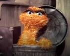 Oscar the Grouch when he was orange in the early days of Sesame Street.  He's in his garbage can, with his head and shoulders exposed and facing forward.  His eyes look very serious.  His left hand is on the edge of the garbage can.