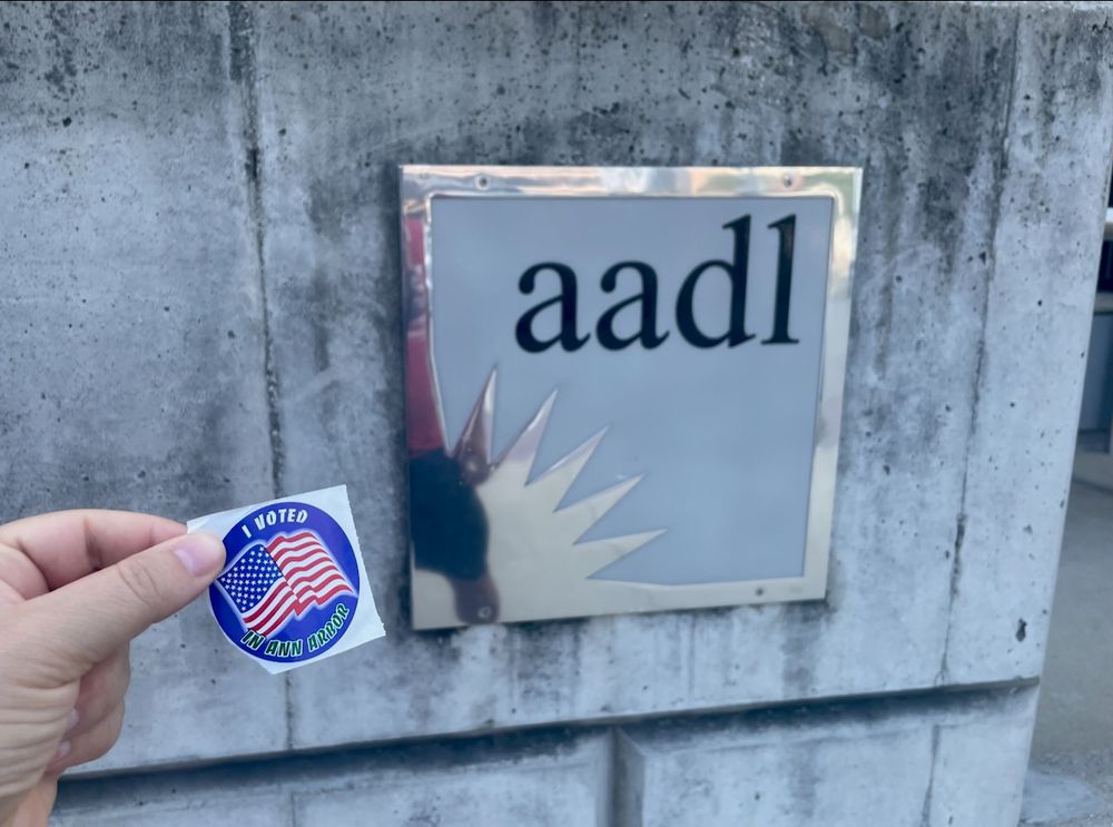 a light-medium skin toned left hand holds up a blue circular sticker with an american flag and text saying, "I voted in Ann Arbor". the hand holds up the sticker in front of the silvered AADL logo on the front of the downtown library branch.