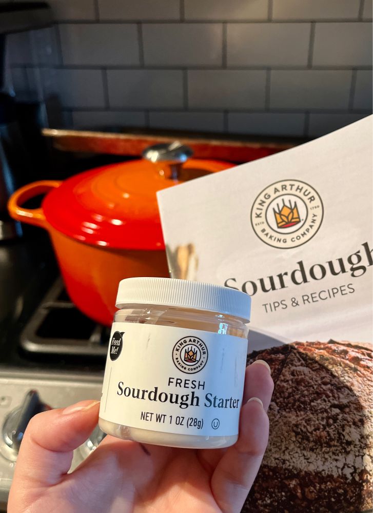 photo of a hand holding a jar labeled fresh sourdough starter with a sourdough tips and recipes booklet and orange dutch oven in the background