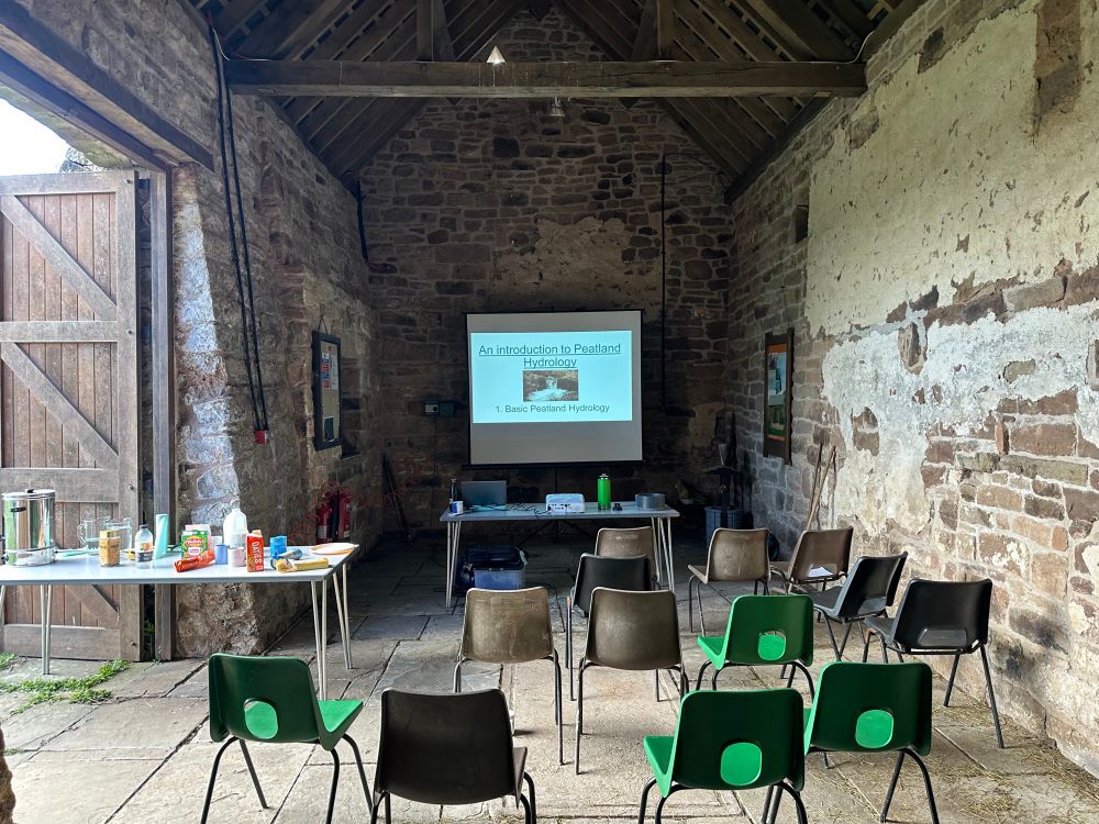 A screen and projector at the front of some chairs in a barn. The slide reads ‘An Introduction to Peatland Hydrology’