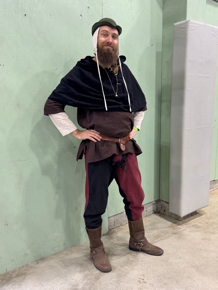 My medievalish outfit for the day: A green bycocket atop a white coif, a smile, a black hood overtop a brown outer tunic and a white inner shirt,  brown leather belt and pouches, alternating red & black trousers, and a pair of brown leather boots.
