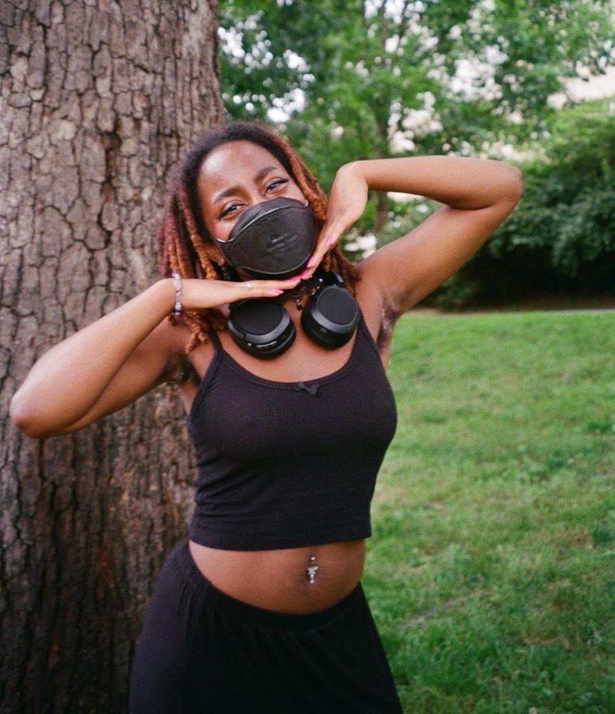 me outdoors in front of a tree. my hair is the same and i’m wearing a black crop tank top, belly piercing, black skirt, and headphones around my neck