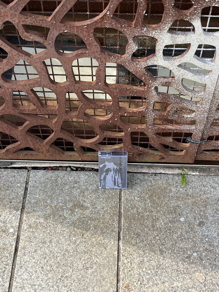A photo of the tape Grains by Andrulian left in public for someone to find. It’s propped against a metal sculpture, location to be determined 