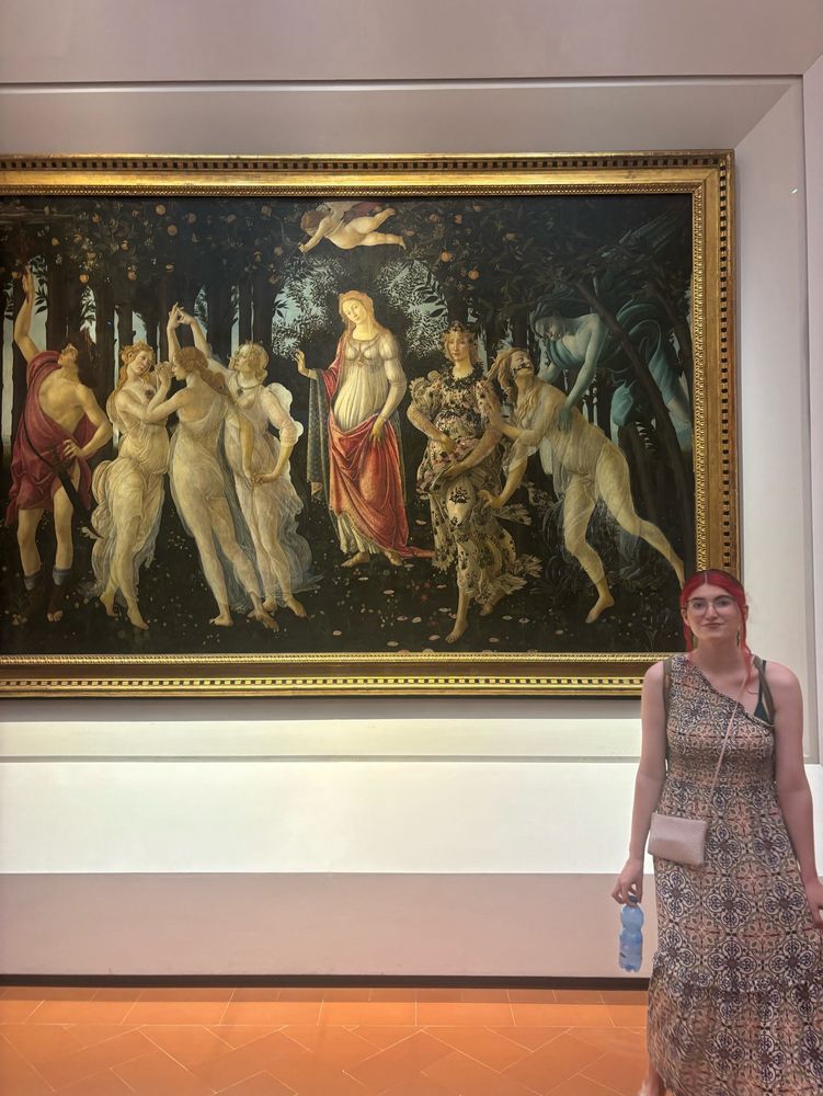 Me, a white woman with dyed red hair wearing a one shoulder, white, creamsicle, and lavender colored ankle length dress in front of Botticelli’s Primavera painting in the Uffizi gallery. I unfortunately look very hot and sweaty and am carrying a half empty bottle of water.
