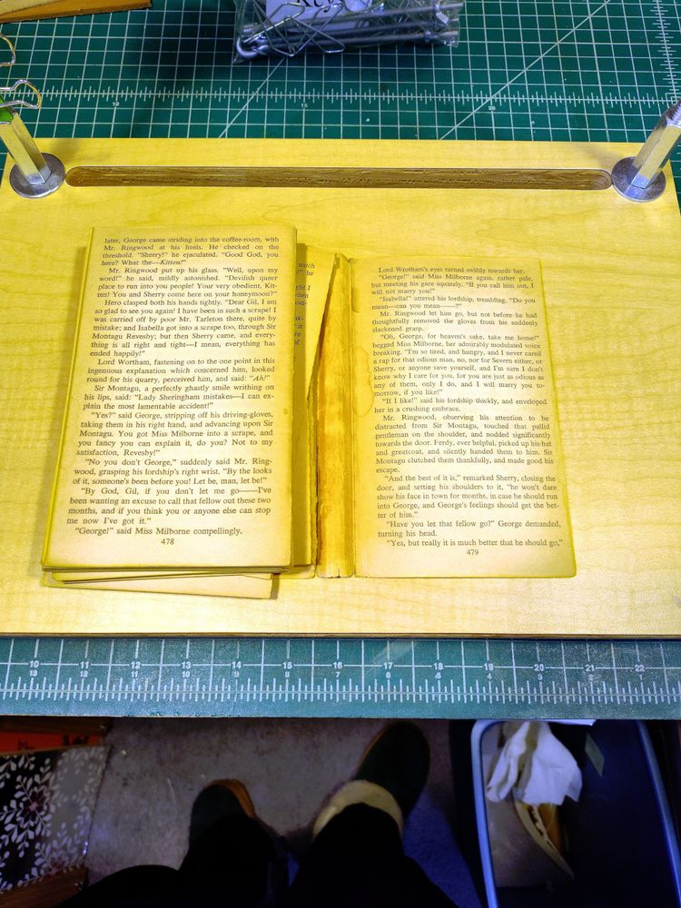 An old paperback book is lying open on a piece of wood resting on a green cutting mat. The book is not looking good, the pages are separating in clumps and the cover is detached.