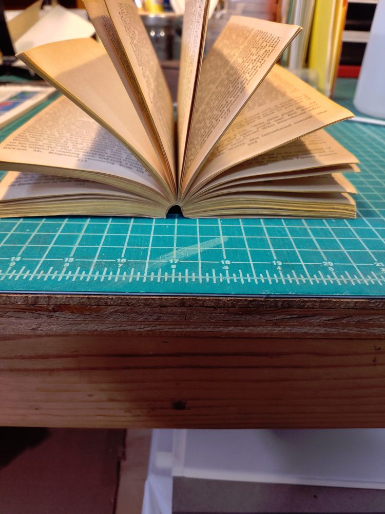 A paperback book is open on a workbench, the photo is focused on the bottom of the book. The spine is curved in a semicircle and the pages are all sticking out nicely.