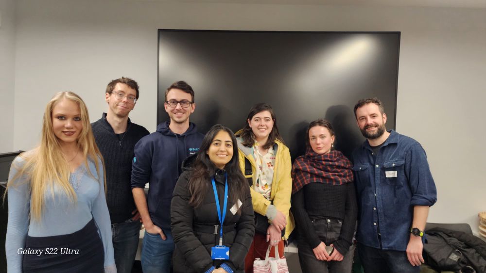 Group of seven speakers, helpers and attendees standing in front of a black TV smiling