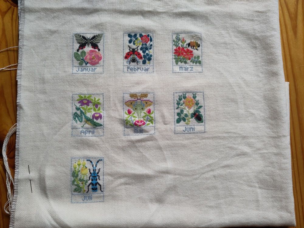 Photo of seven cross stitched rectangles with colorful flowers and different bugs and insects. Underneath each rectangle is the name of a month in German from "Januar" to "Juli". 