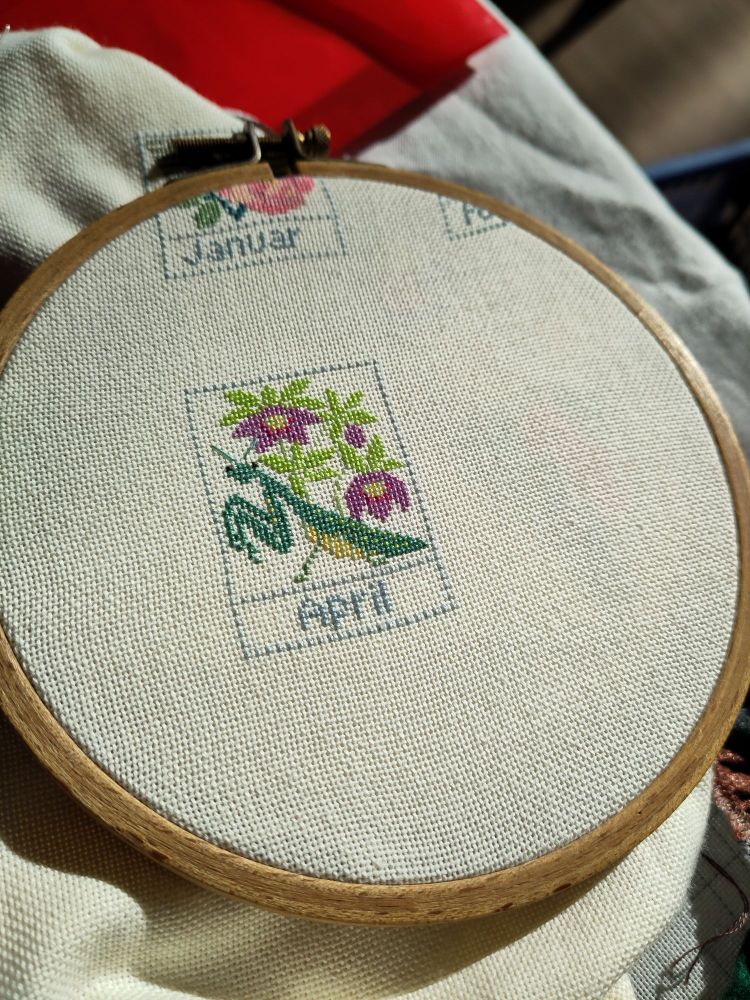 Photo of a cross stitch in a wooden hoop. It's a small rectangle depicting a praying mantis in front of purple flowers. At the bottom is the text "April".