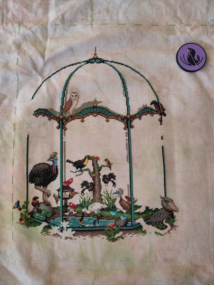 Photo of a cross stitch in progress. Lots of colorful birds in a giant ornate aviary on a creamy fabric with brown and green spots.