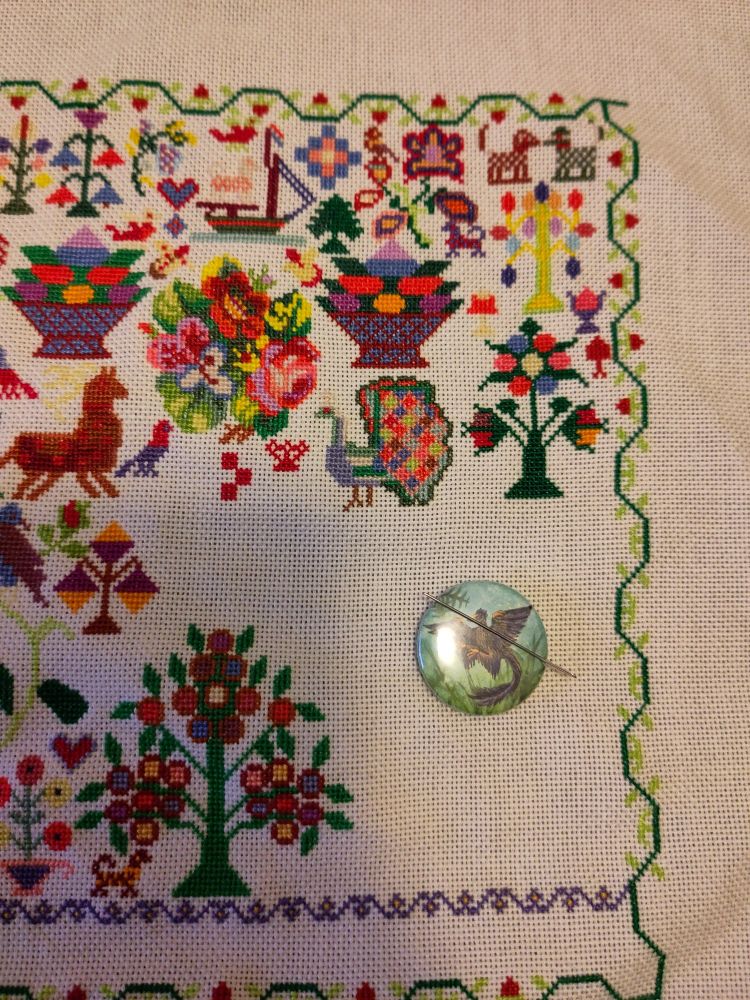 Photo of a cross stitch in progress in a wooden hoop. It's a colorful sampler with a strawberry border full of traditional and funky motifs, like two small dogs, a flower, a big berry bowl, brightly colored trees with fruit, and a motley peacock.