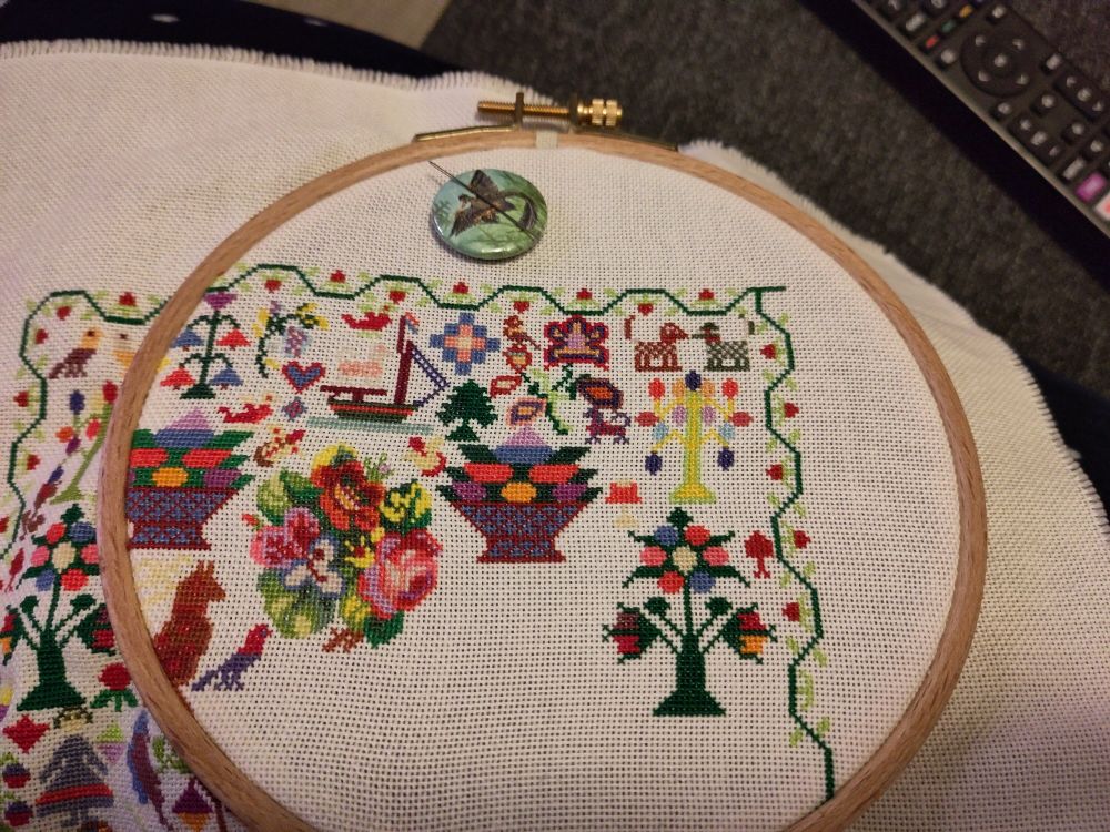 Photo of a cross stitch in progress in a wooden hoop. It's a colorful sampler with a strawberry border full of traditional and funky motifs, like two small dogs, a flower, a big berry bowl, and brightly colored trees with oblong and round fruit in various colors.