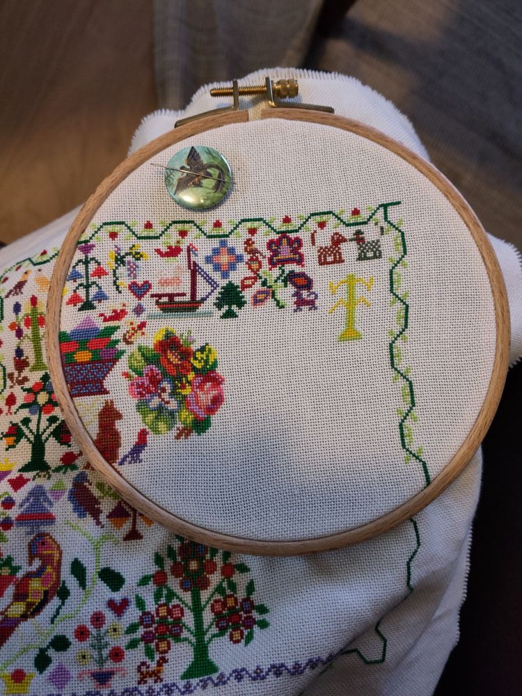Photo of a cross stitch in progress in a wooden hoop. It's a colorful sampler with a strawberry border full of traditional and funky motifs, like two small dogs, a flower. The border is brought down further on the right side and there are parts of a new, light green and yellow tree.