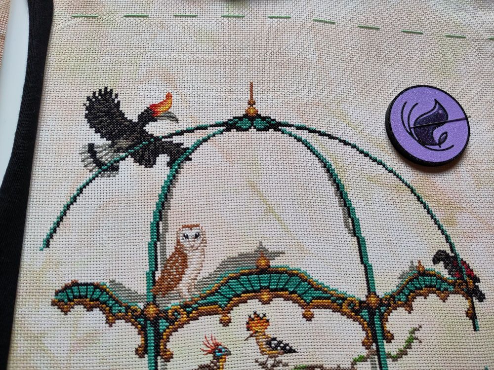 Photo of a cross stitch in progress. On marbled light brown fabric, the top part of an ornate aviary with a couple of colorful birds. In the top left corner, a hornbill in flight.
