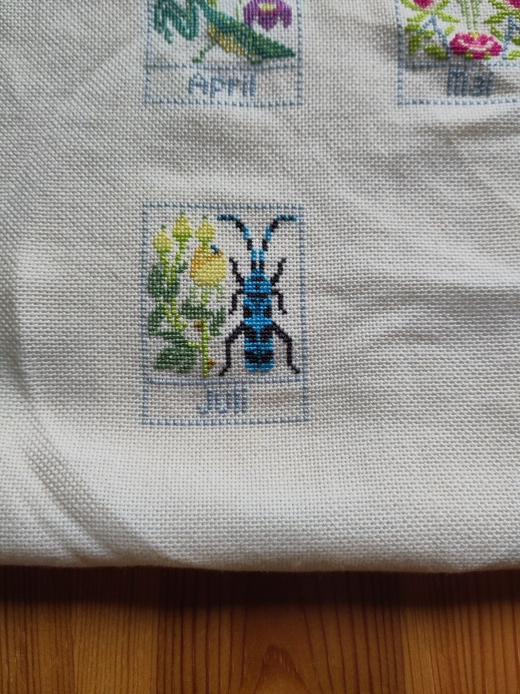 Photo of a cross stitch: a rectangle with a yellow flower and a big black and blue bug. Underneath, the text "Juli".